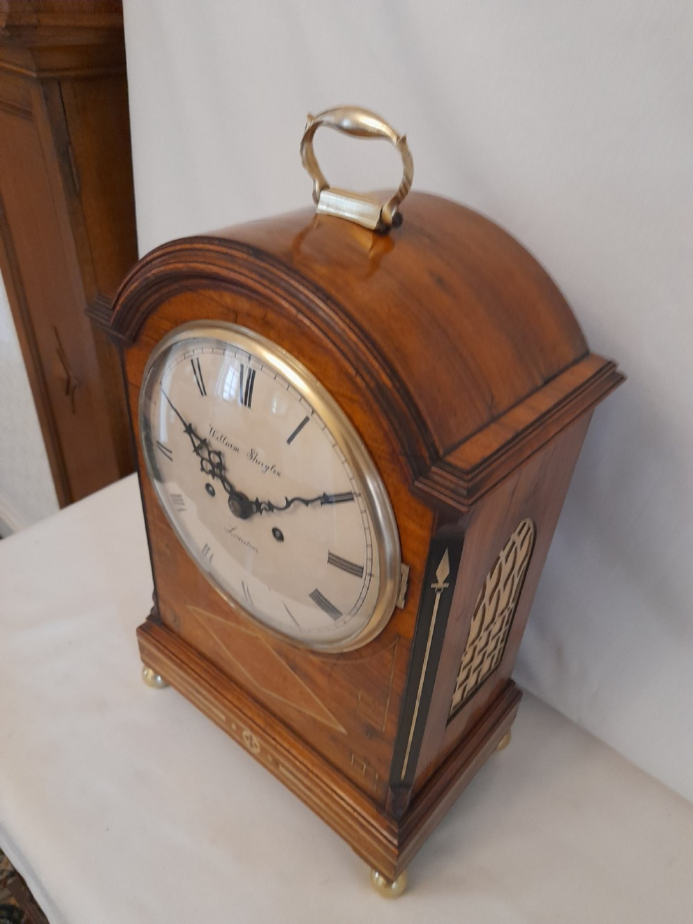 A Regency period rosewood bracket clock featuring geometric brass inlay, arched top with brass handle, gothic side frets, circular dial, and brass ball feet. View VIII