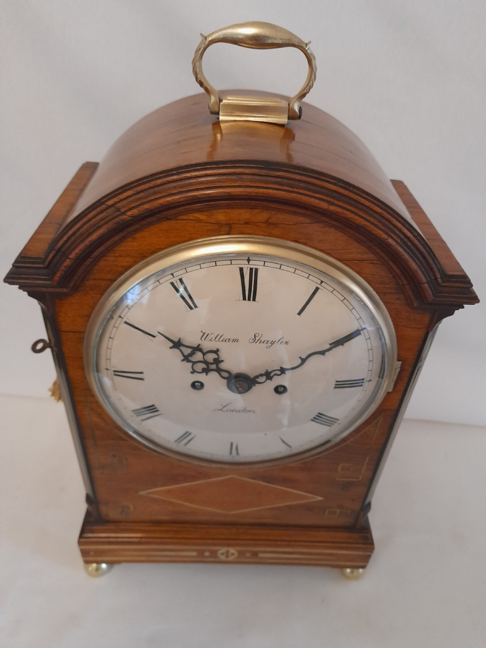 A Regency period rosewood bracket clock featuring geometric brass inlay, arched top with brass handle, gothic side frets, circular dial, and brass ball feet. View IX