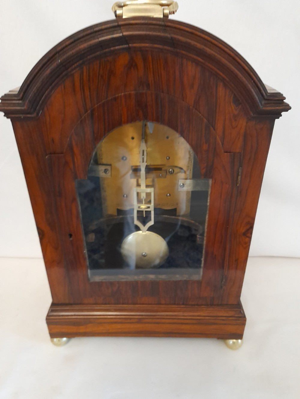 A Regency period rosewood bracket clock featuring geometric brass inlay, arched top with brass handle, gothic side frets, circular dial, and brass ball feet. View X