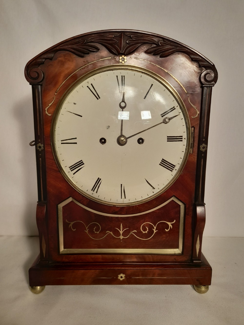 A Georgian mahogany bracket clock featuring brass inlay decoration, carved foliage cresting, and a white circular dial with Roman numerals.