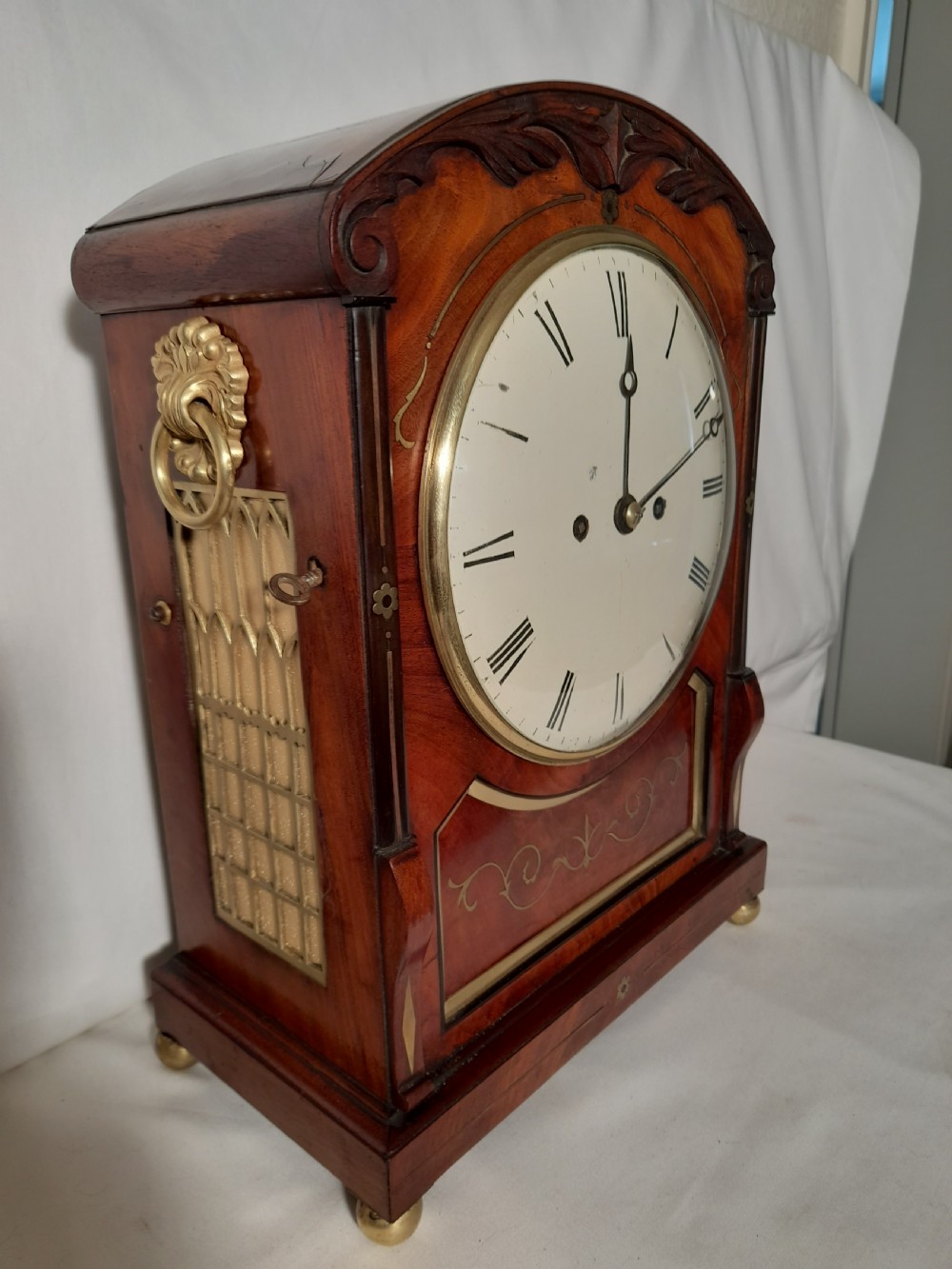 A Georgian mahogany bracket clock featuring brass inlay decoration, carved foliage cresting, and a white circular dial with Roman numerals. View III
