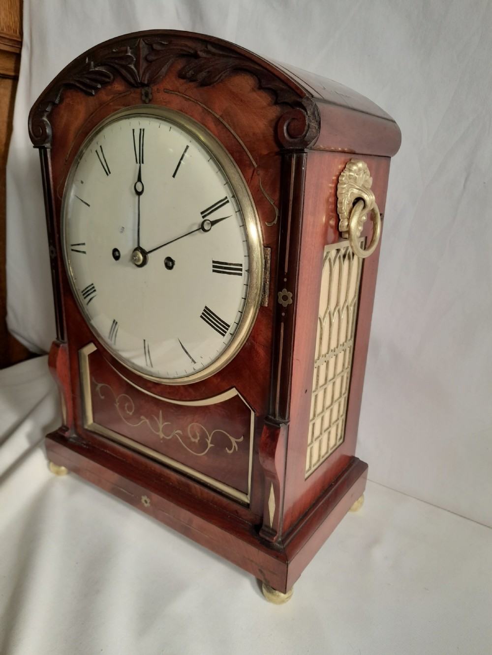 A Georgian mahogany bracket clock featuring brass inlay decoration, carved foliage cresting, and a white circular dial with Roman numerals. View V