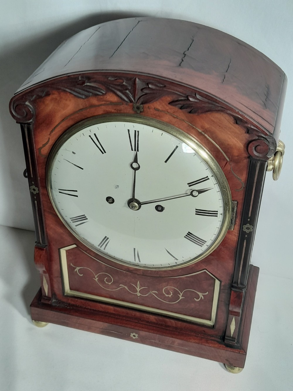 A Georgian mahogany bracket clock featuring brass inlay decoration, carved foliage cresting, and a white circular dial with Roman numerals. View VII