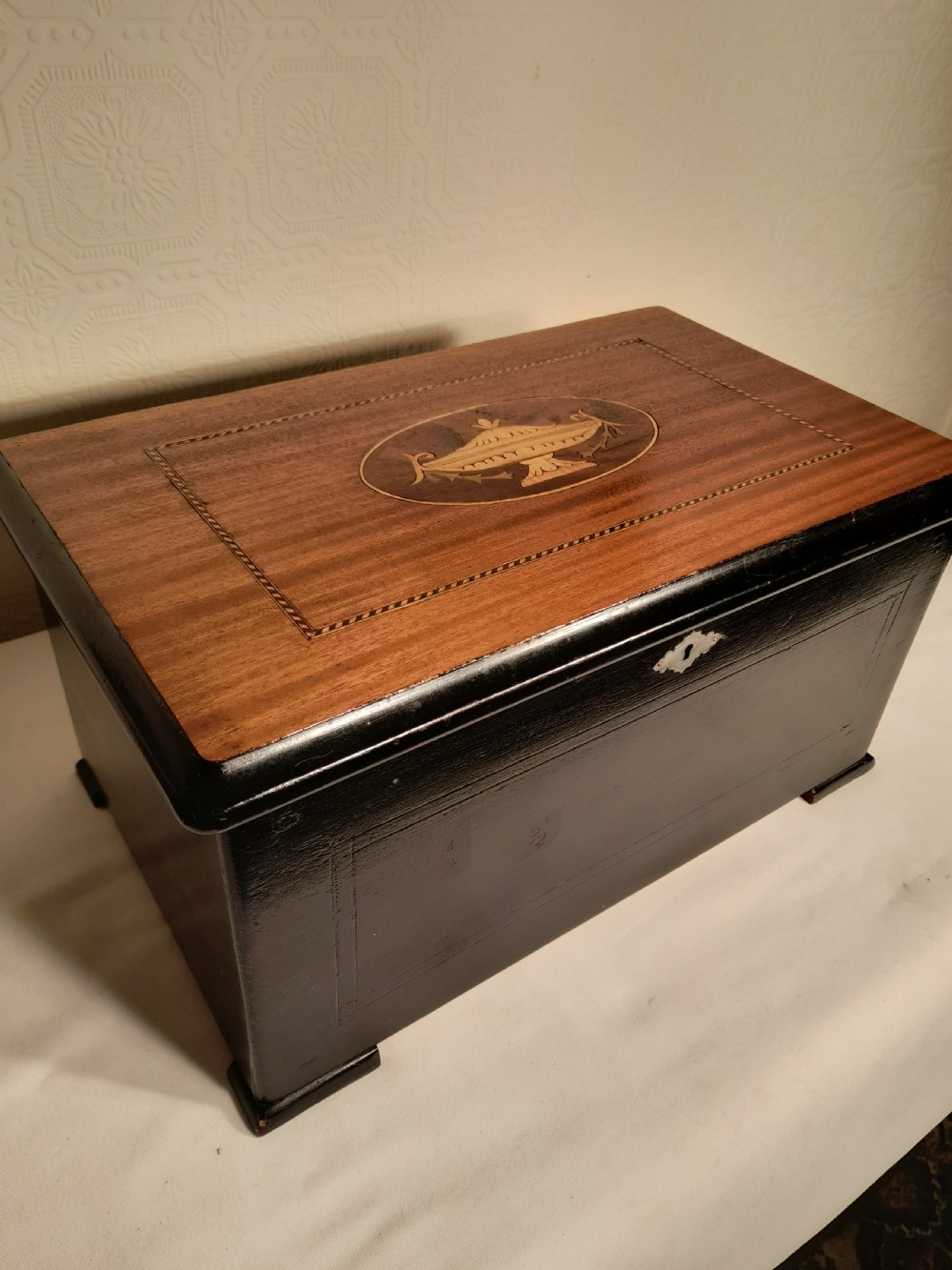 An antique wooden music box with an ebonised lower case, a mahogany hinged lid featuring a marquetry urn inlay, and a central keyhole.