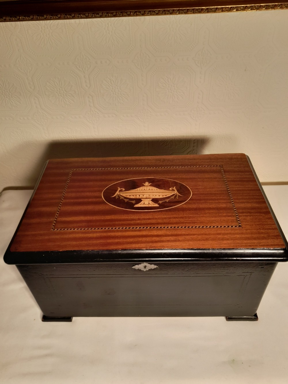 An antique wooden music box with an ebonised lower case, a mahogany hinged lid featuring a marquetry urn inlay, and a central keyhole. View IV