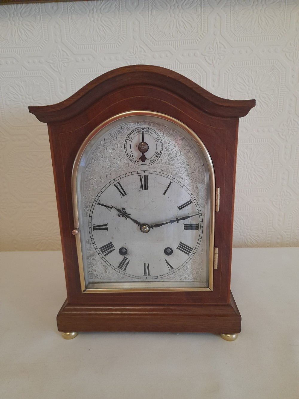 An arched mahogany wooden bracket clock with a silvered brass dial displaying Roman numerals and a small regulating dial at the top.