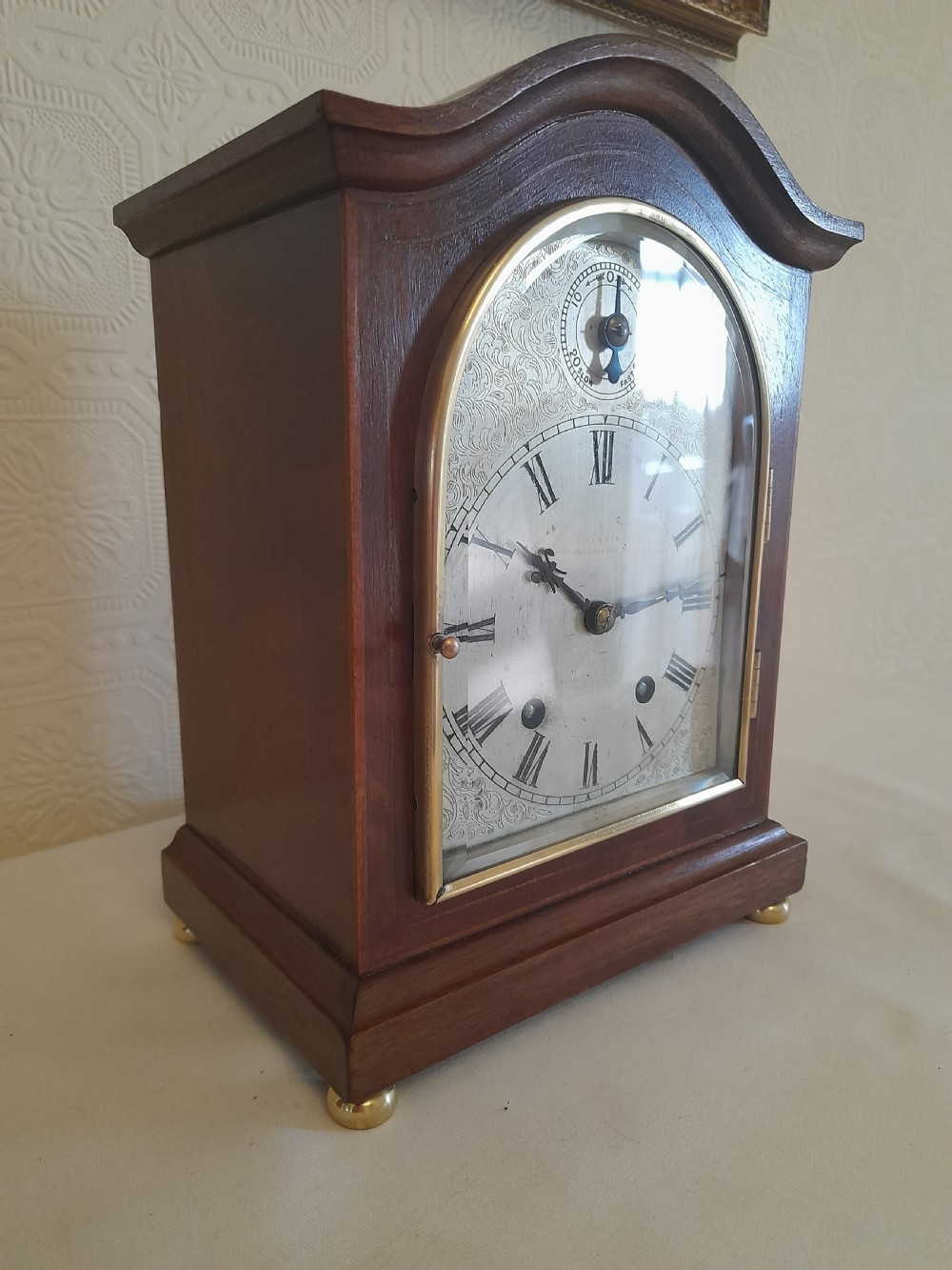 An arched mahogany wooden bracket clock with a silvered brass dial displaying Roman numerals and a small regulating dial at the top. View III