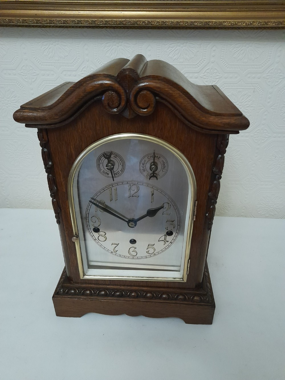An Edwardian carved oak bracket clock featuring a silvered dial, brass bezel, arch top scroll moulding, and Westminster chime regulation sub-dials.