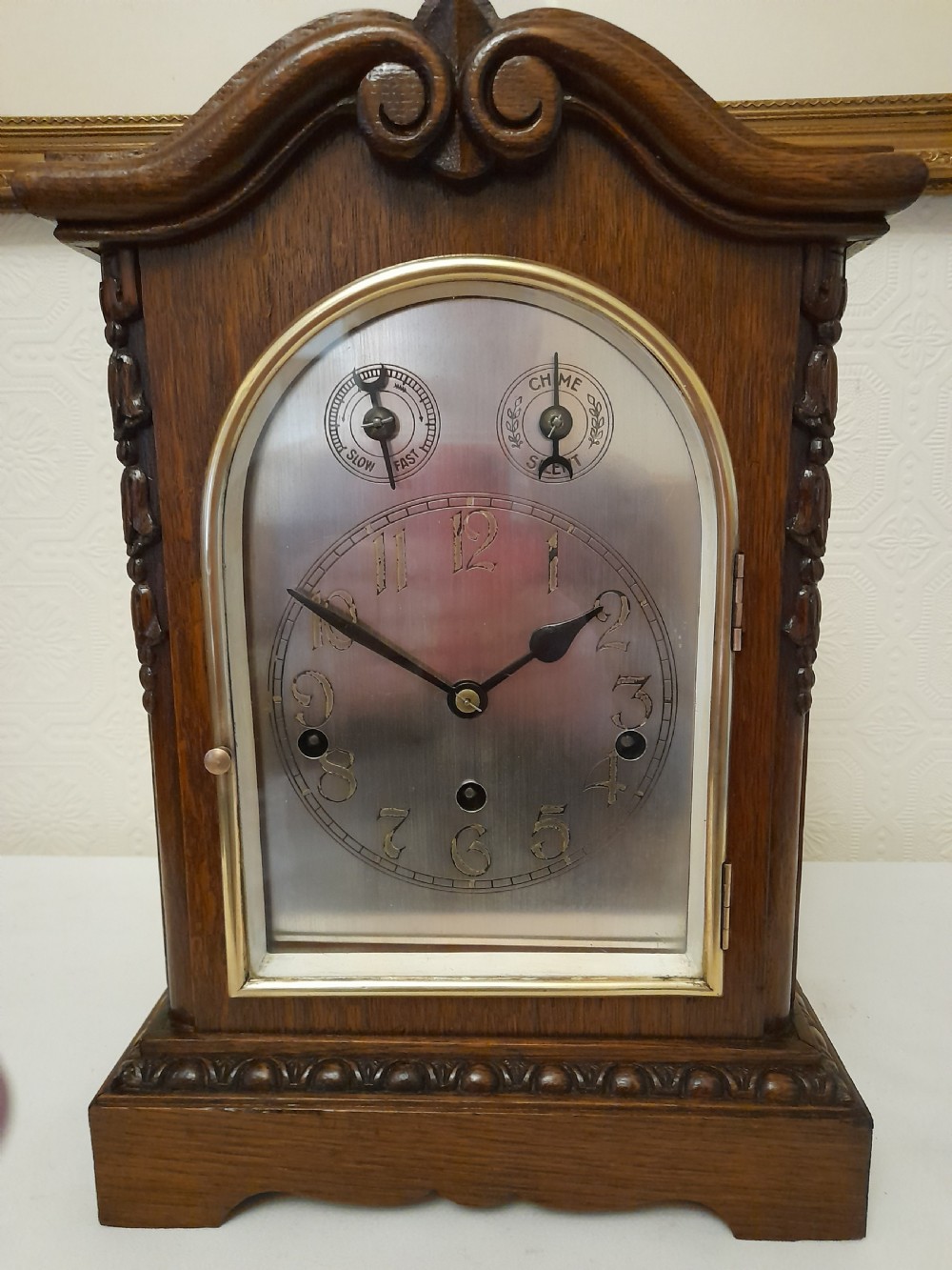 An Edwardian carved oak bracket clock featuring a silvered dial, brass bezel, arch top scroll moulding, and Westminster chime regulation sub-dials. View II