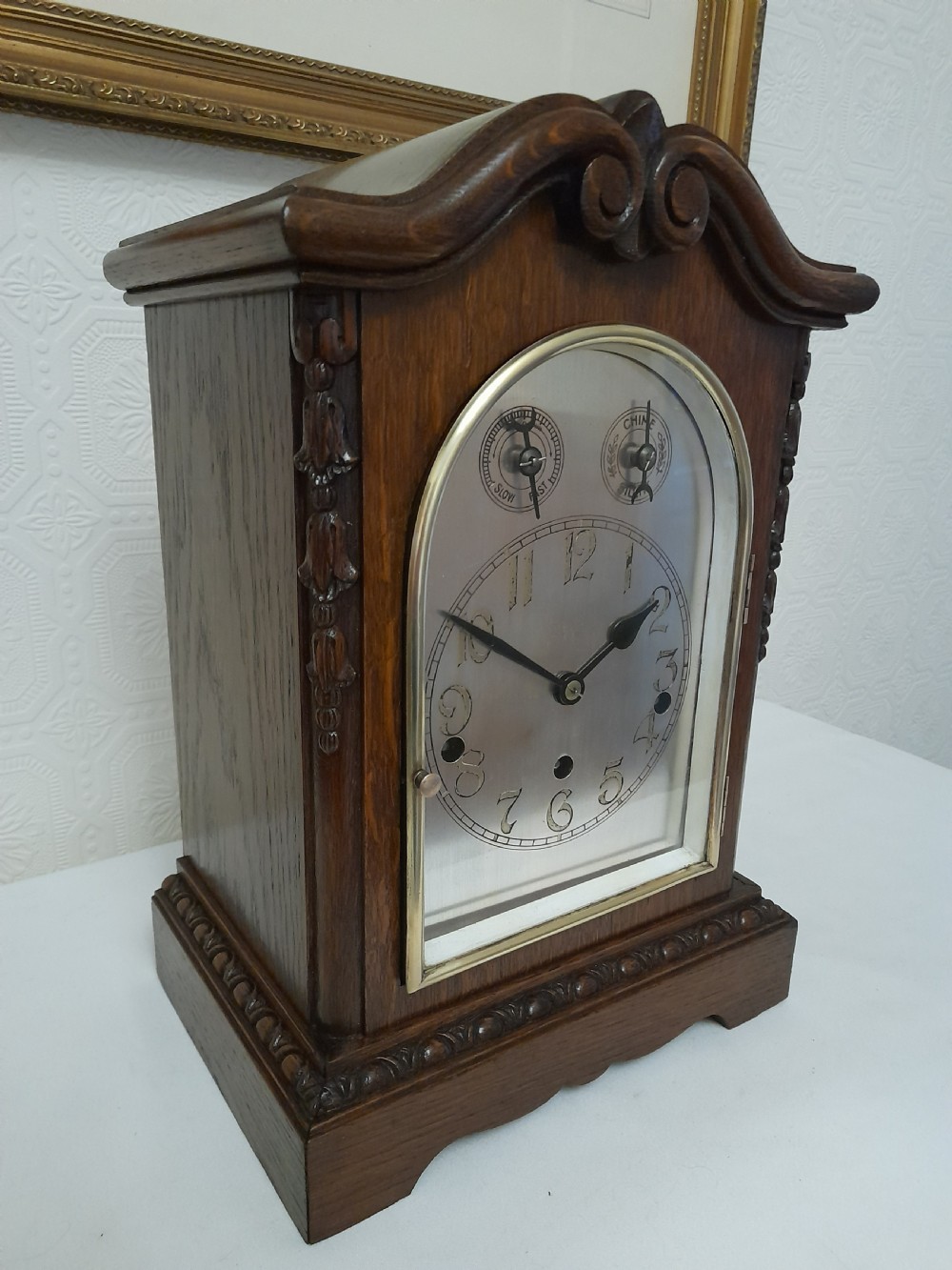 An Edwardian carved oak bracket clock featuring a silvered dial, brass bezel, arch top scroll moulding, and Westminster chime regulation sub-dials. View V