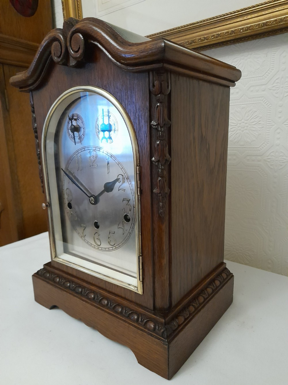An Edwardian carved oak bracket clock featuring a silvered dial, brass bezel, arch top scroll moulding, and Westminster chime regulation sub-dials. View VI