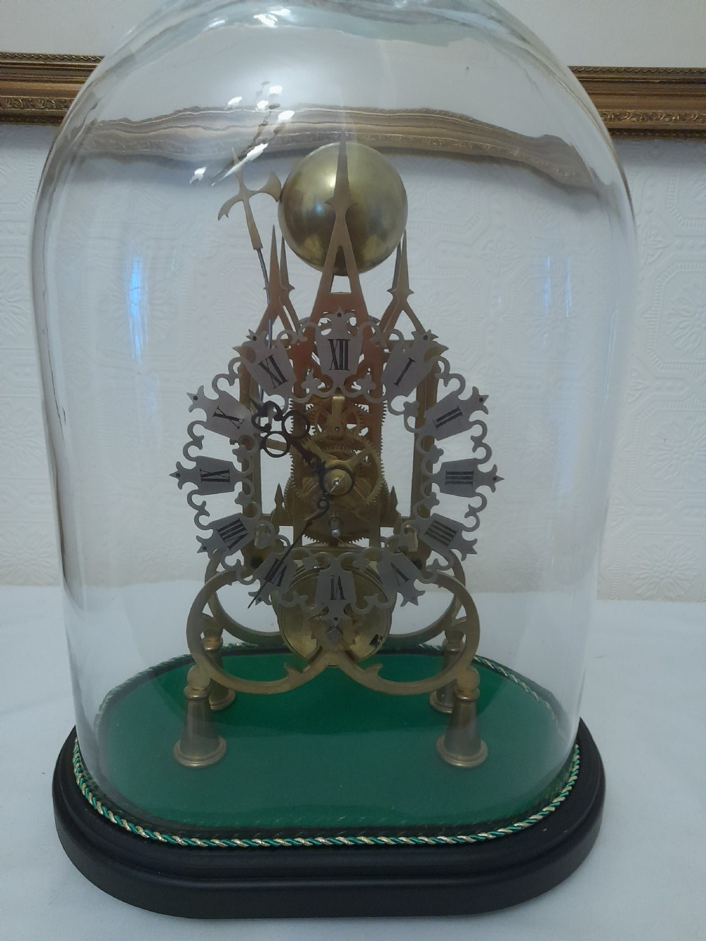 A Victorian brass skeleton clock featuring exposed internal gears and a silvered Roman numeral dial, encased under a clear glass dome on an oval wooden base.