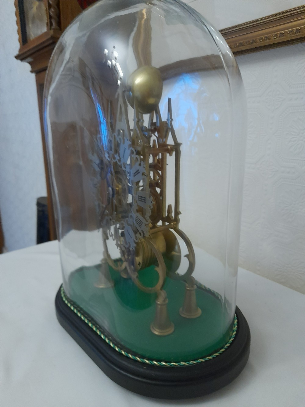 A Victorian brass skeleton clock featuring exposed internal gears and a silvered Roman numeral dial, encased under a clear glass dome on an oval wooden base. View II