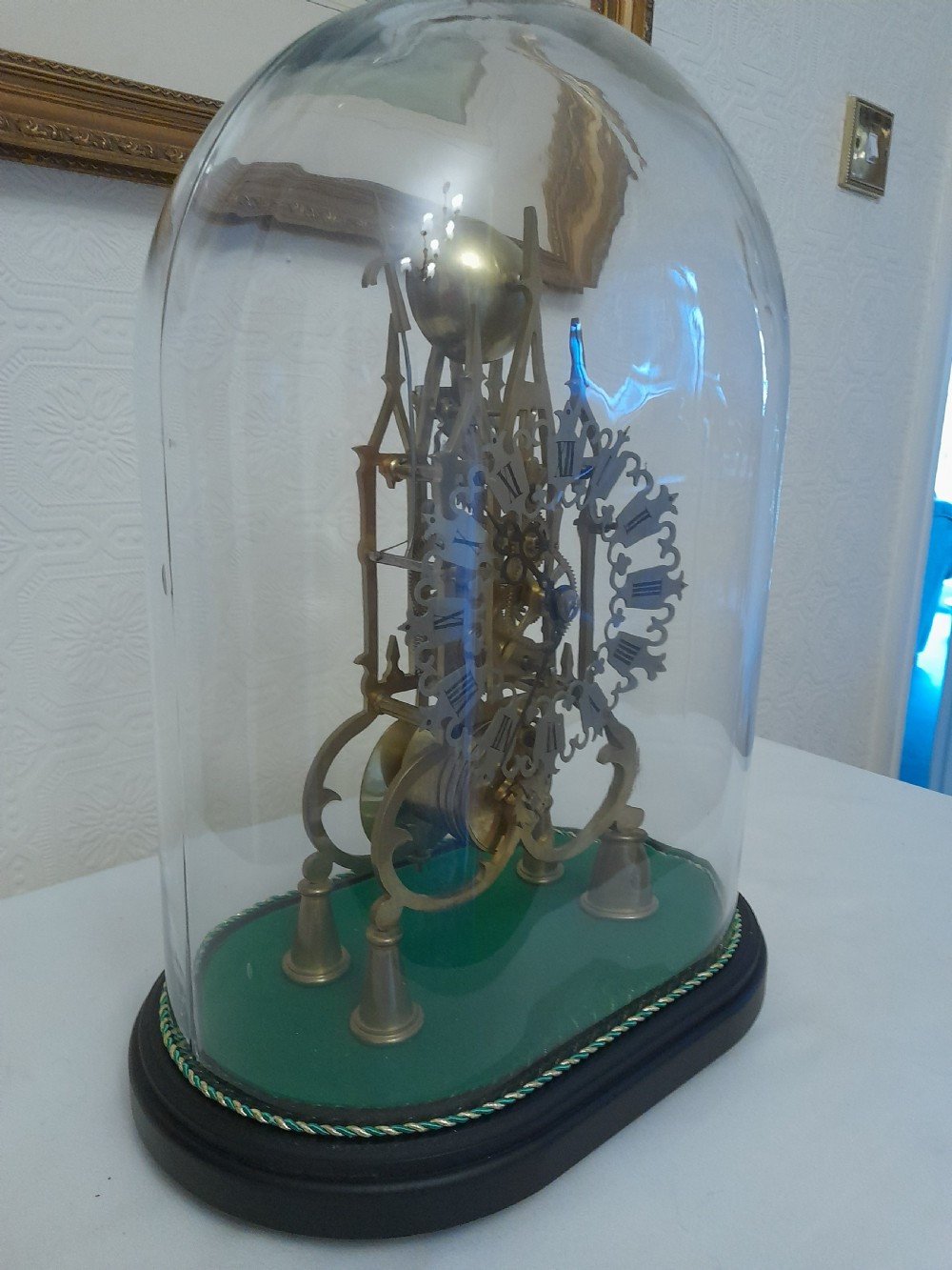 A Victorian brass skeleton clock featuring exposed internal gears and a silvered Roman numeral dial, encased under a clear glass dome on an oval wooden base. View III