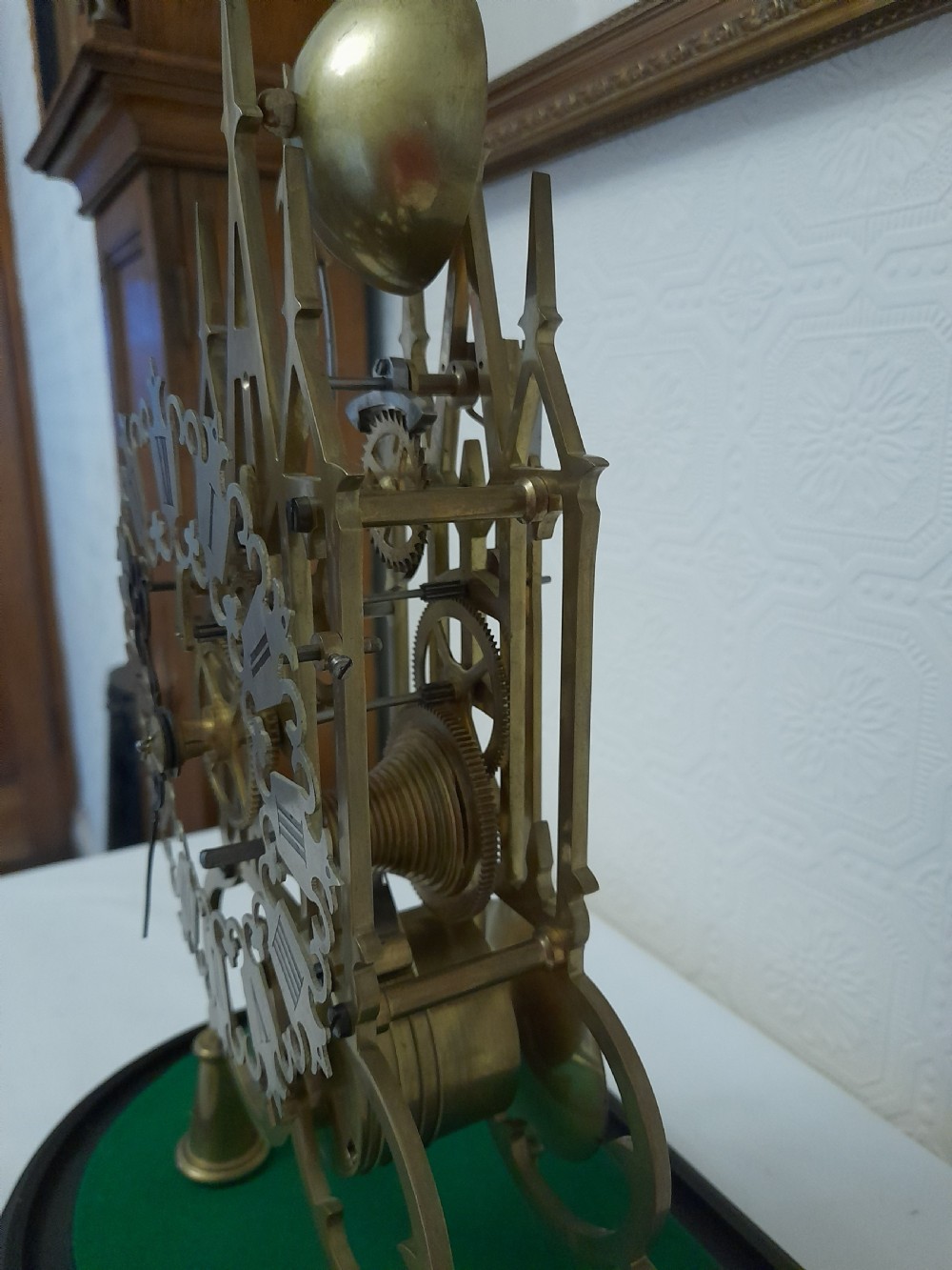 A Victorian brass skeleton clock featuring exposed internal gears and a silvered Roman numeral dial, encased under a clear glass dome on an oval wooden base. View V