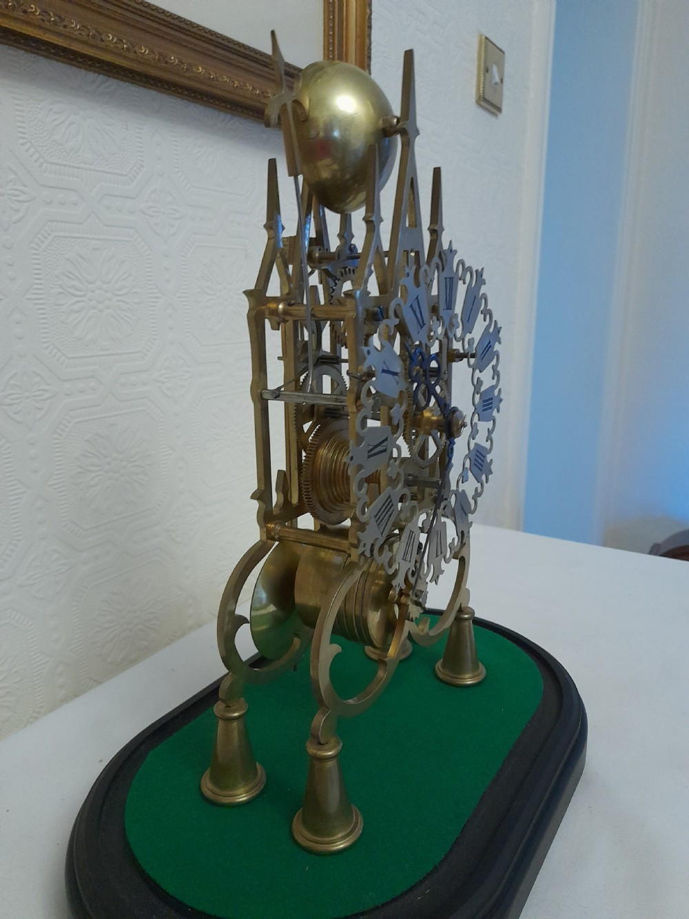 A Victorian brass skeleton clock featuring exposed internal gears and a silvered Roman numeral dial, encased under a clear glass dome on an oval wooden base. View VI