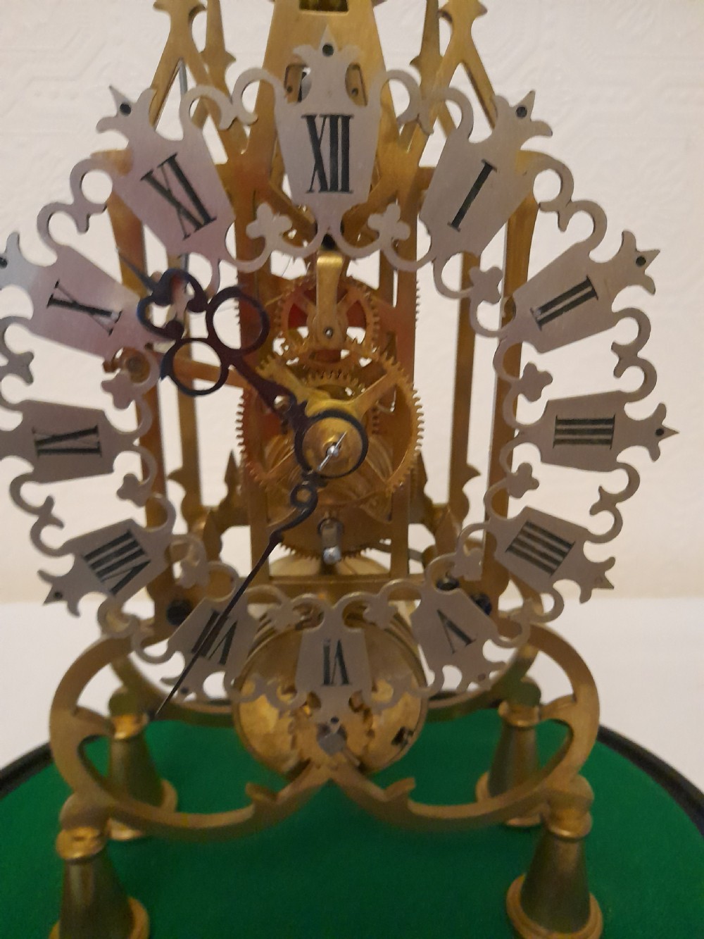 A Victorian brass skeleton clock featuring exposed internal gears and a silvered Roman numeral dial, encased under a clear glass dome on an oval wooden base. View VIII