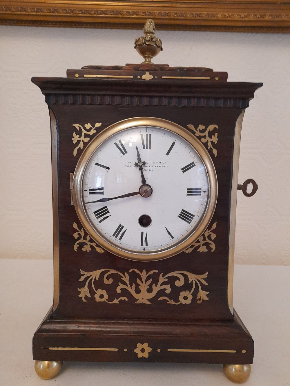 A Regency dark wood mantel clock featuring floral brass inlays, a white circular dial with Roman numerals, brass ball feet, and a top finial.