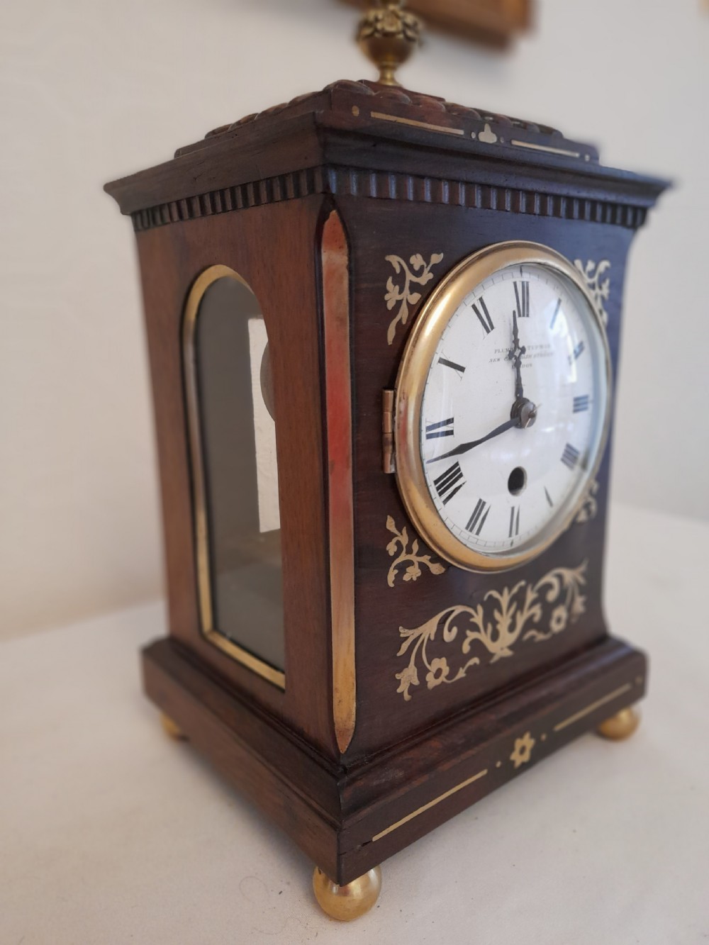 A Regency dark wood mantel clock featuring floral brass inlays, a white circular dial with Roman numerals, brass ball feet, and a top finial. View II