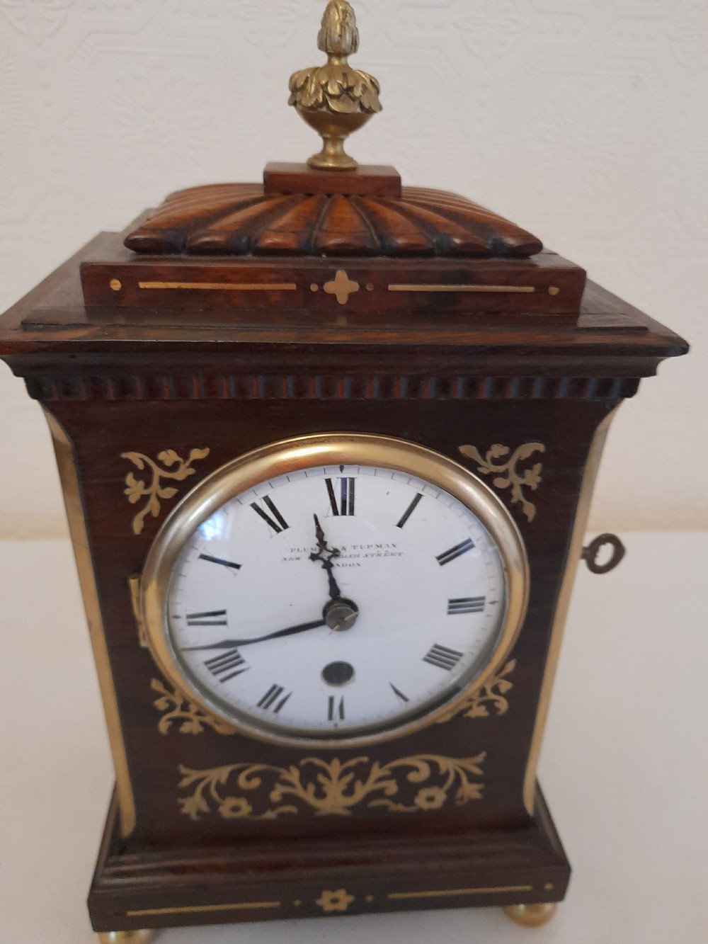 A Regency dark wood mantel clock featuring floral brass inlays, a white circular dial with Roman numerals, brass ball feet, and a top finial. View VI