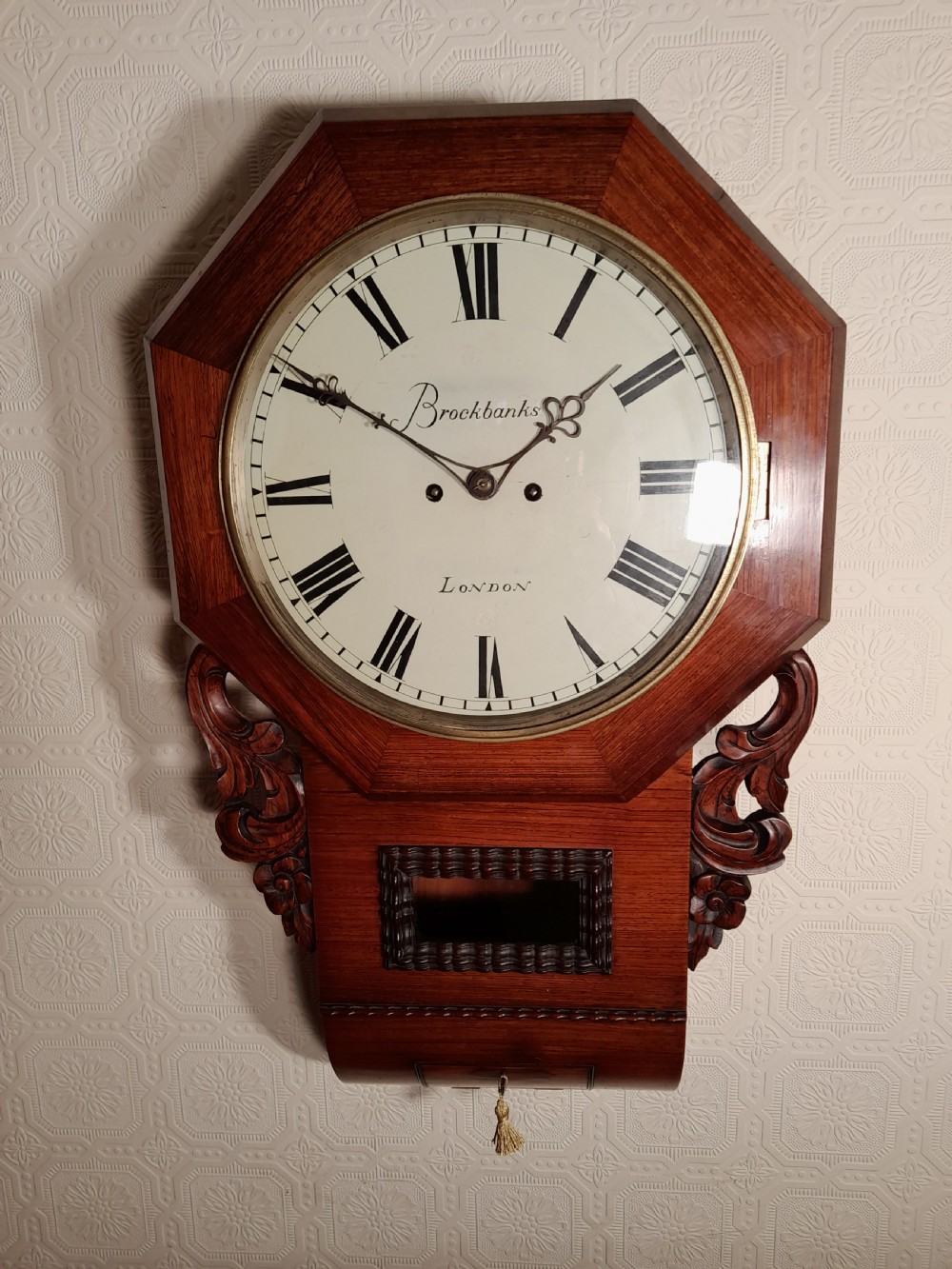 An antique mahogany drop-dial wall clock featuring an octagonal bezel frame, cream dial with Roman numerals, signature Brockbanks London, and carved side scrolls.
