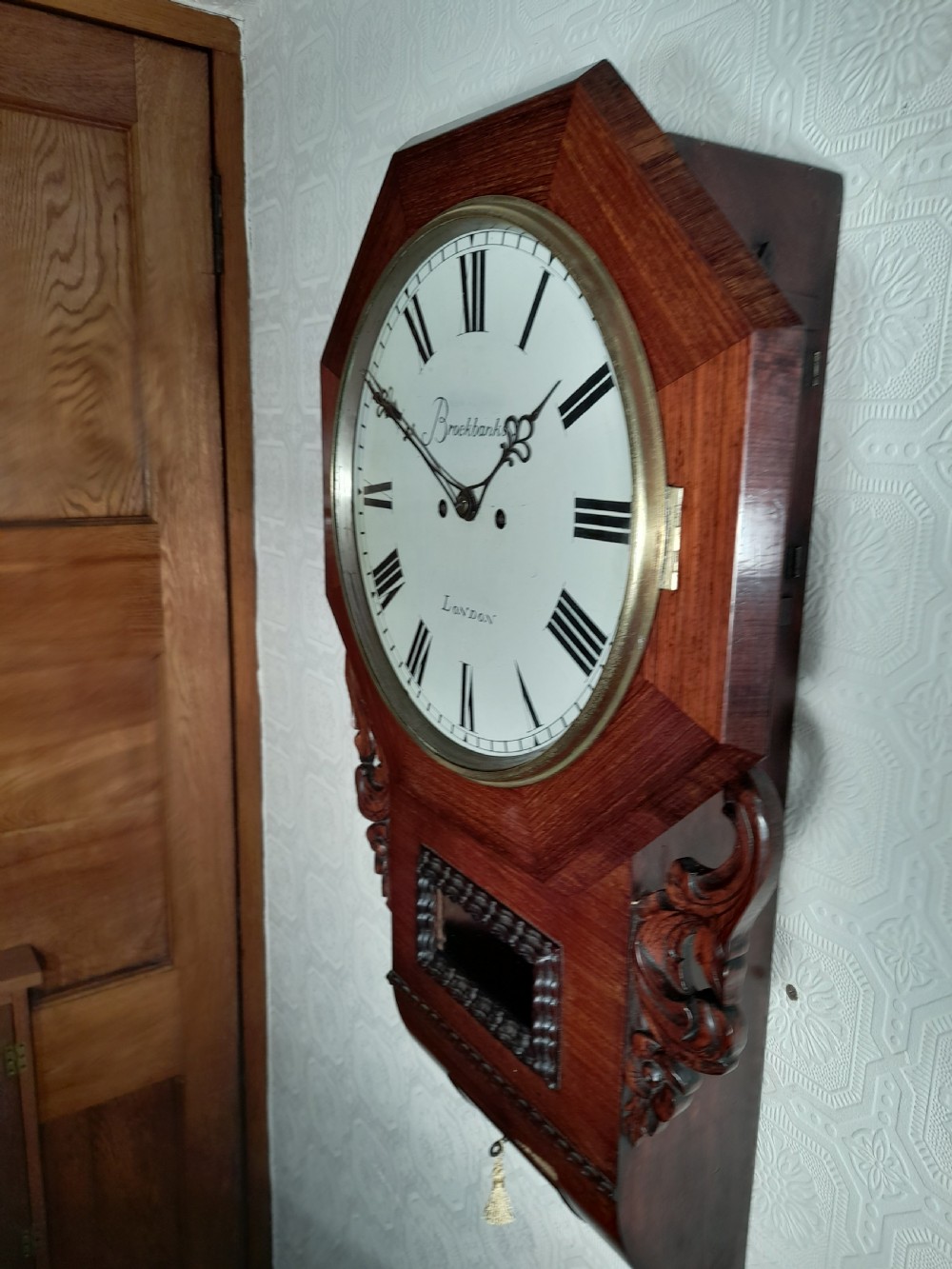 An antique mahogany drop-dial wall clock featuring an octagonal bezel frame, cream dial with Roman numerals, signature Brockbanks London, and carved side scrolls. View VII