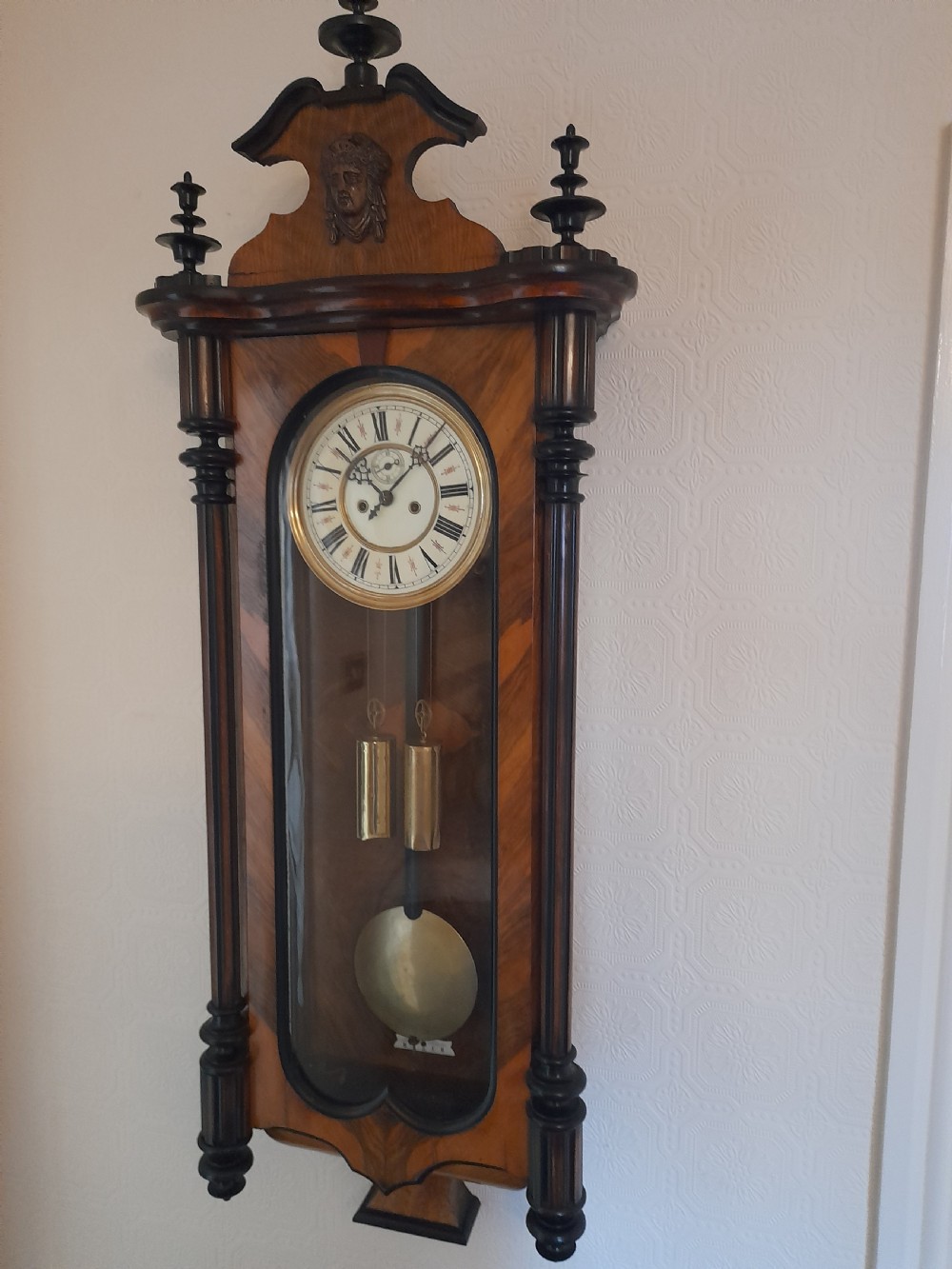 A dark walnut-cased Gustav Becker twin-weight Vienna regulator wall clock featuring turned side columns, carved cresting, twin brass weights, and a white enamel Roman numeral dial.