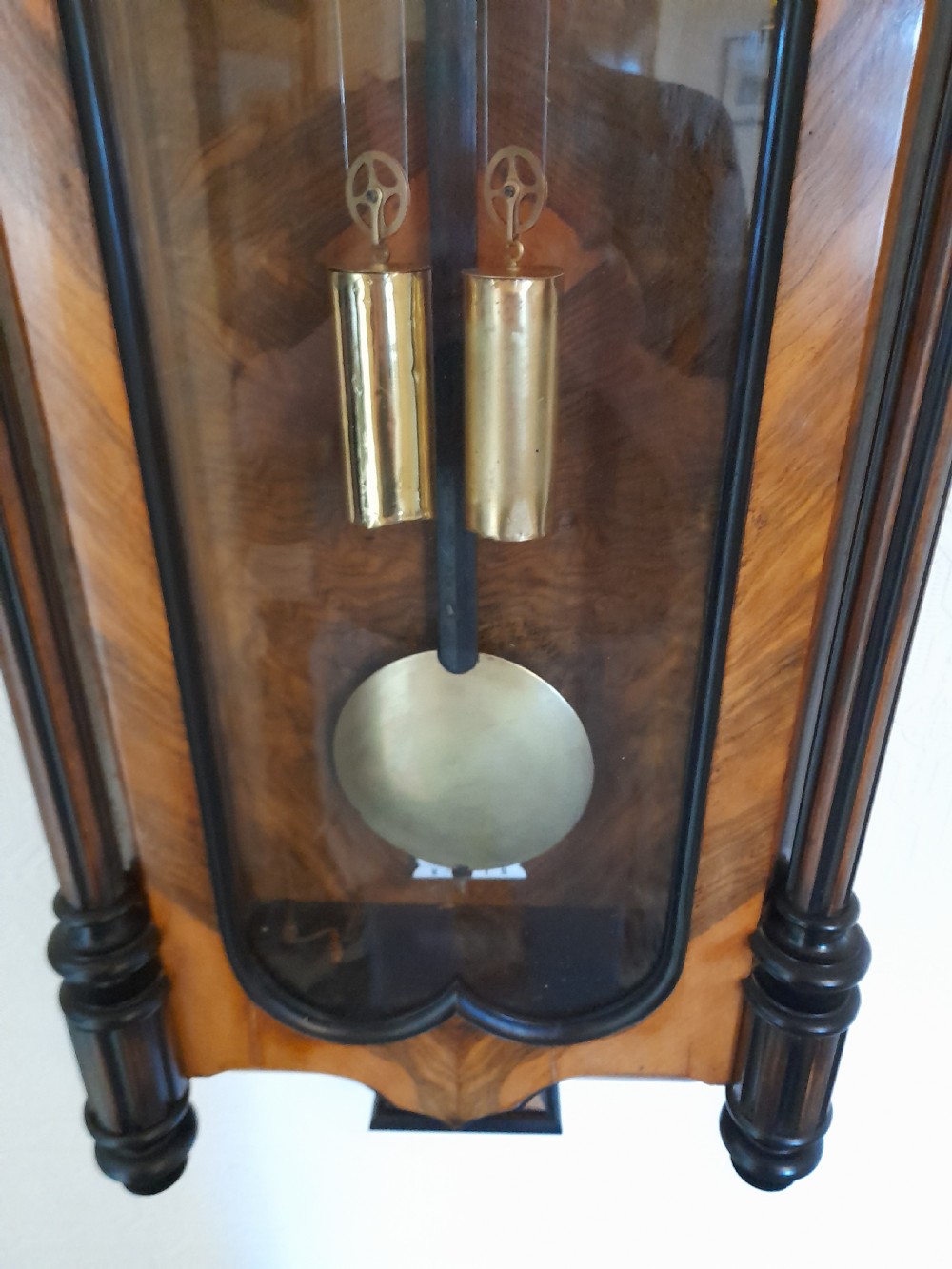 A dark walnut-cased Gustav Becker twin-weight Vienna regulator wall clock featuring turned side columns, carved cresting, twin brass weights, and a white enamel Roman numeral dial. View V