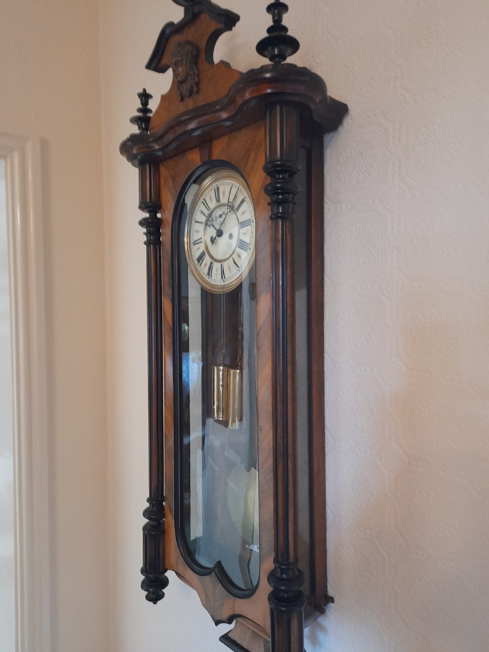 A dark walnut-cased Gustav Becker twin-weight Vienna regulator wall clock featuring turned side columns, carved cresting, twin brass weights, and a white enamel Roman numeral dial. View VI