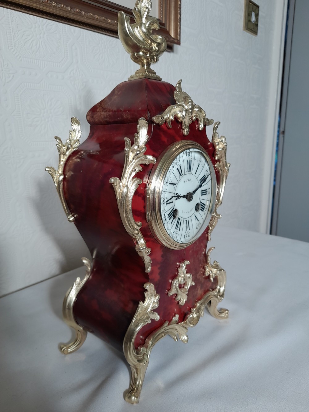 A French Rococo Revival style mantel clock featuring crimson tortoiseshell veneering, ornate gilt bronze mounts, and a white enamel dial with Roman numerals. View V