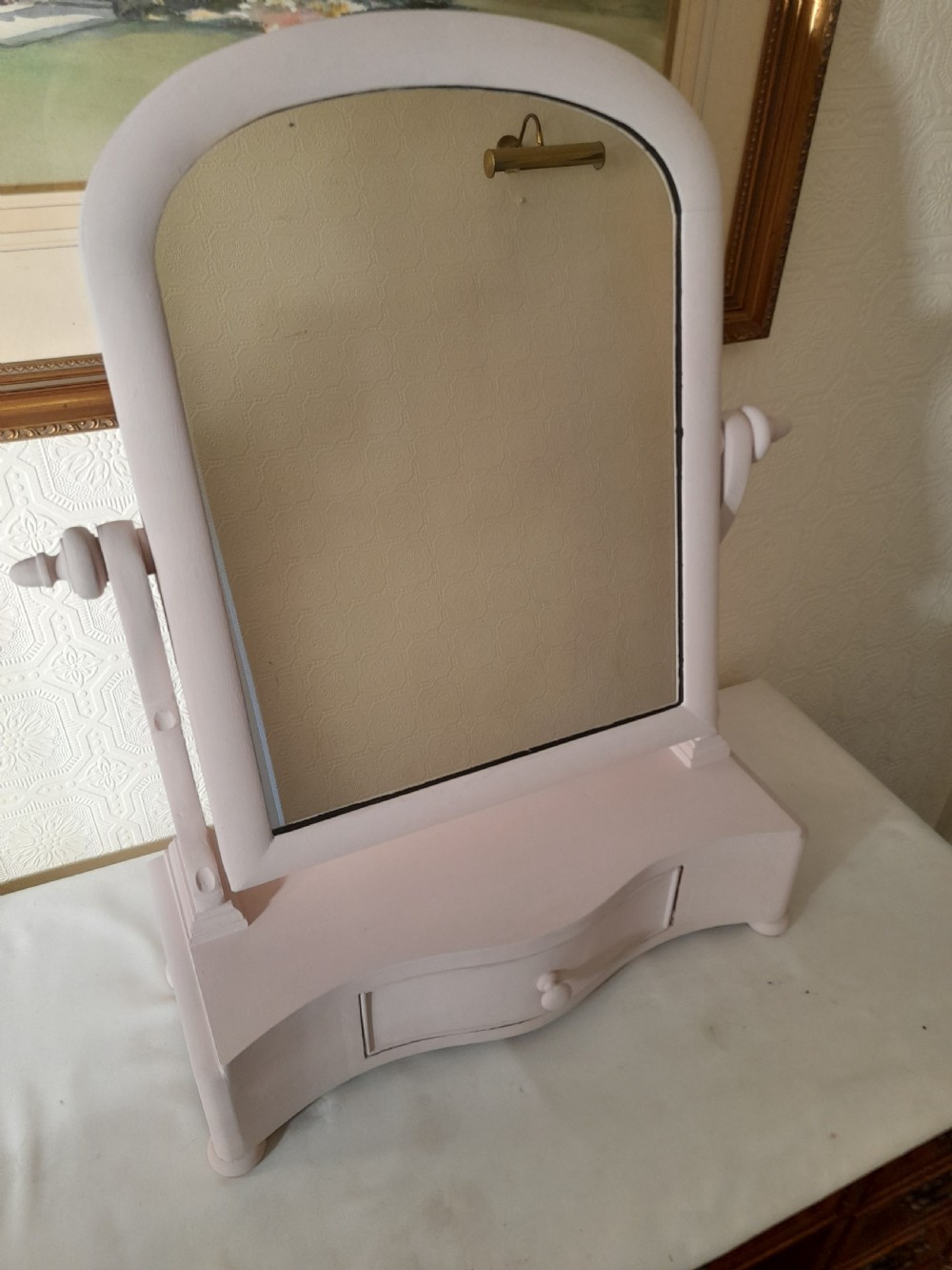 A pale pink painted wooden dressing table mirror featuring an arched tilting glass frame, side turned support pins, and a single drawer in the base plinth.