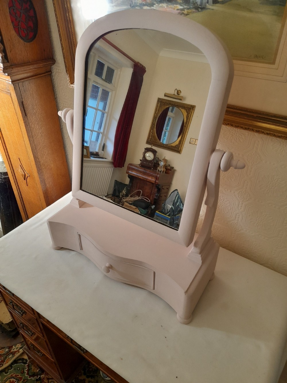 A pale pink painted wooden dressing table mirror featuring an arched tilting glass frame, side turned support pins, and a single drawer in the base plinth. View II