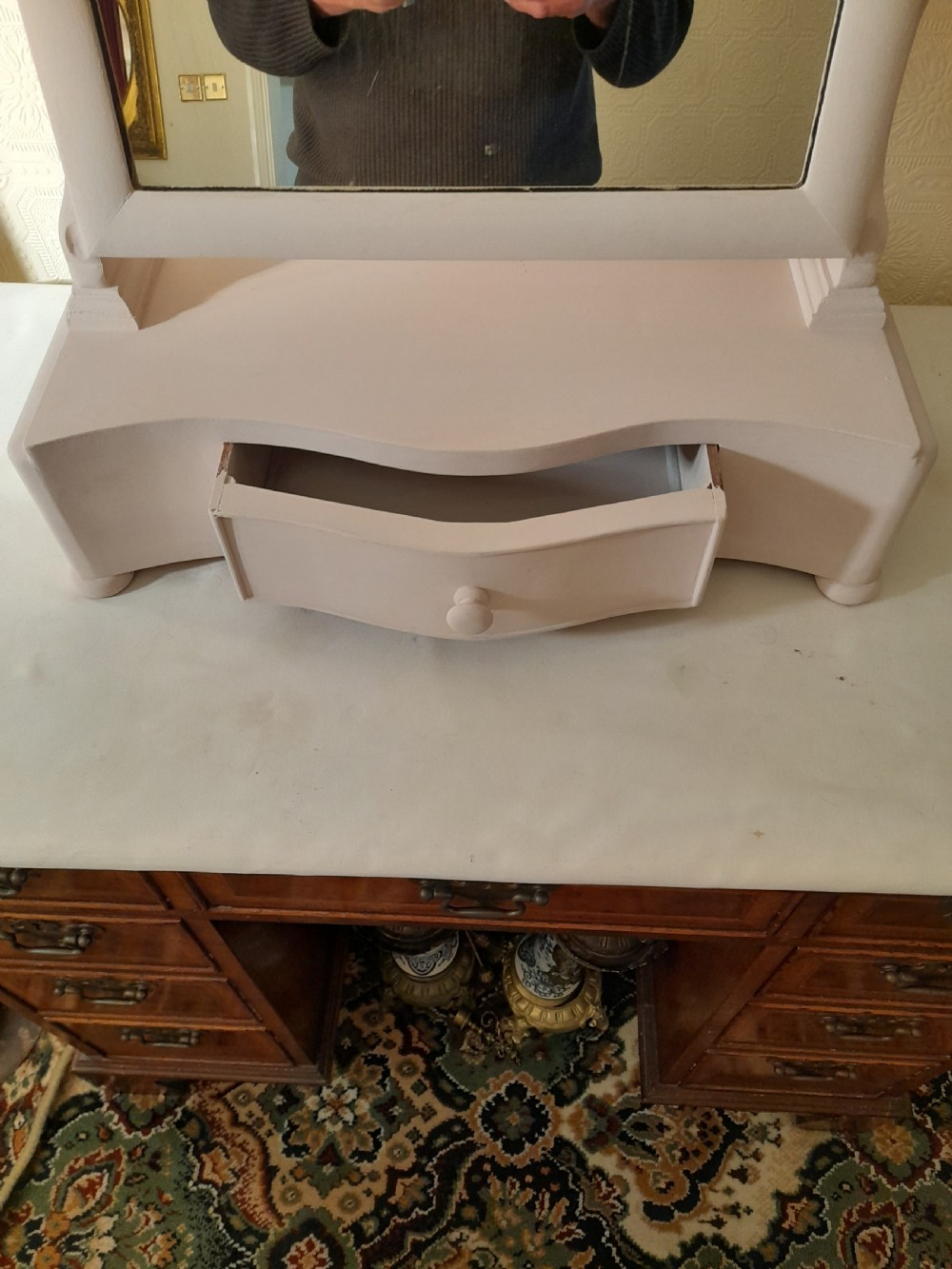 A pale pink painted wooden dressing table mirror featuring an arched tilting glass frame, side turned support pins, and a single drawer in the base plinth. View III