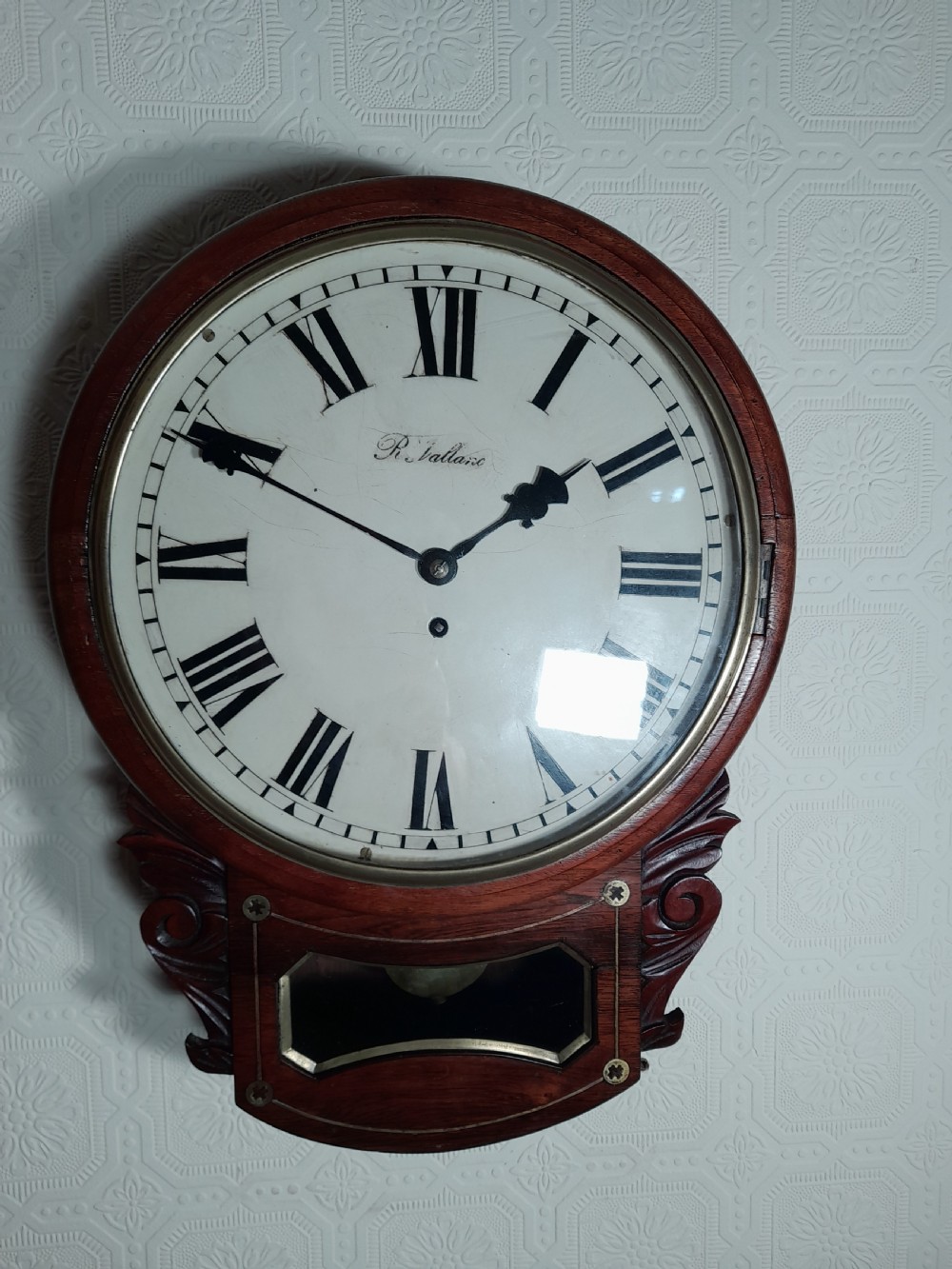 A Victorian rosewood drop-dial wall clock with Roman numerals on a white face, brass accents, and a lower glazed pendulum window. View VI