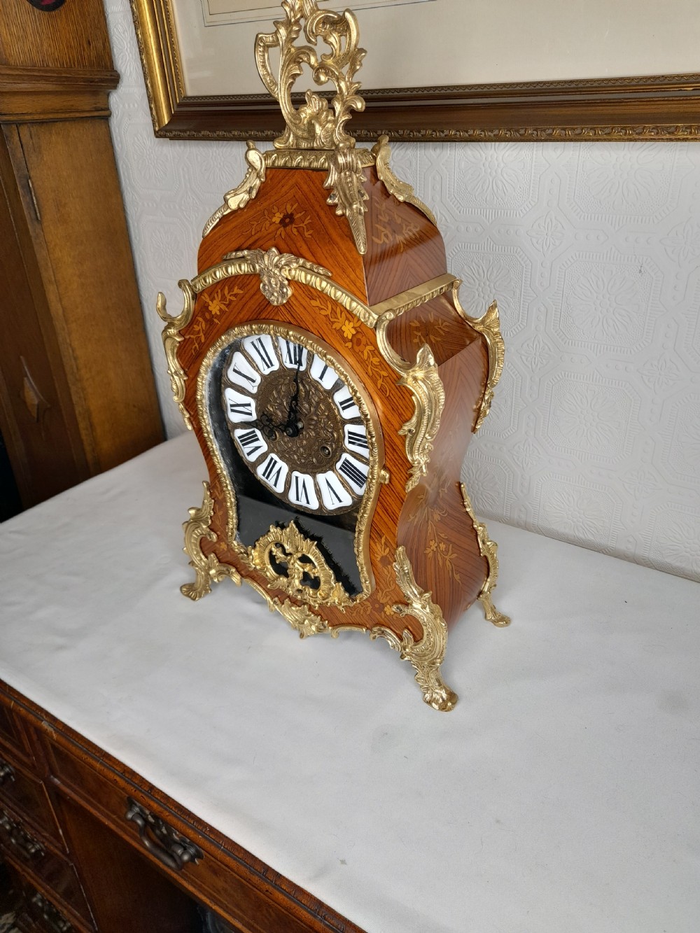 A Louis XIV style bracket clock featuring floral wood marquetry, ornate gilt-bronze mounts, and a dial with white enamel Roman numeral cartouches. View VI