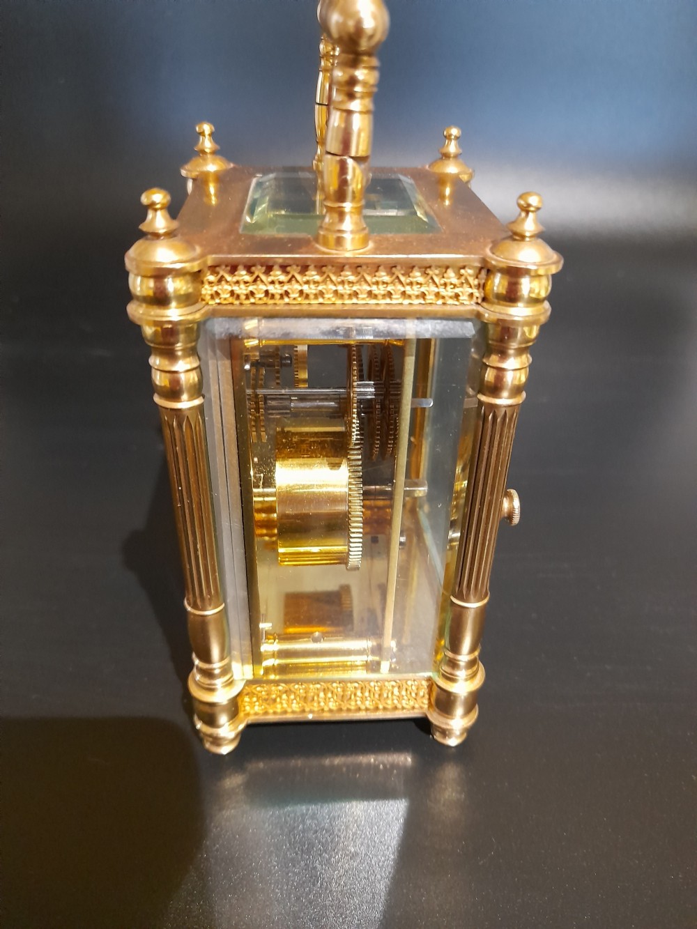 A gilded brass carriage clock featuring clear glass side panels, turned corner columns, a folding top handle, and a white enamel dial with Arabic numerals. View III