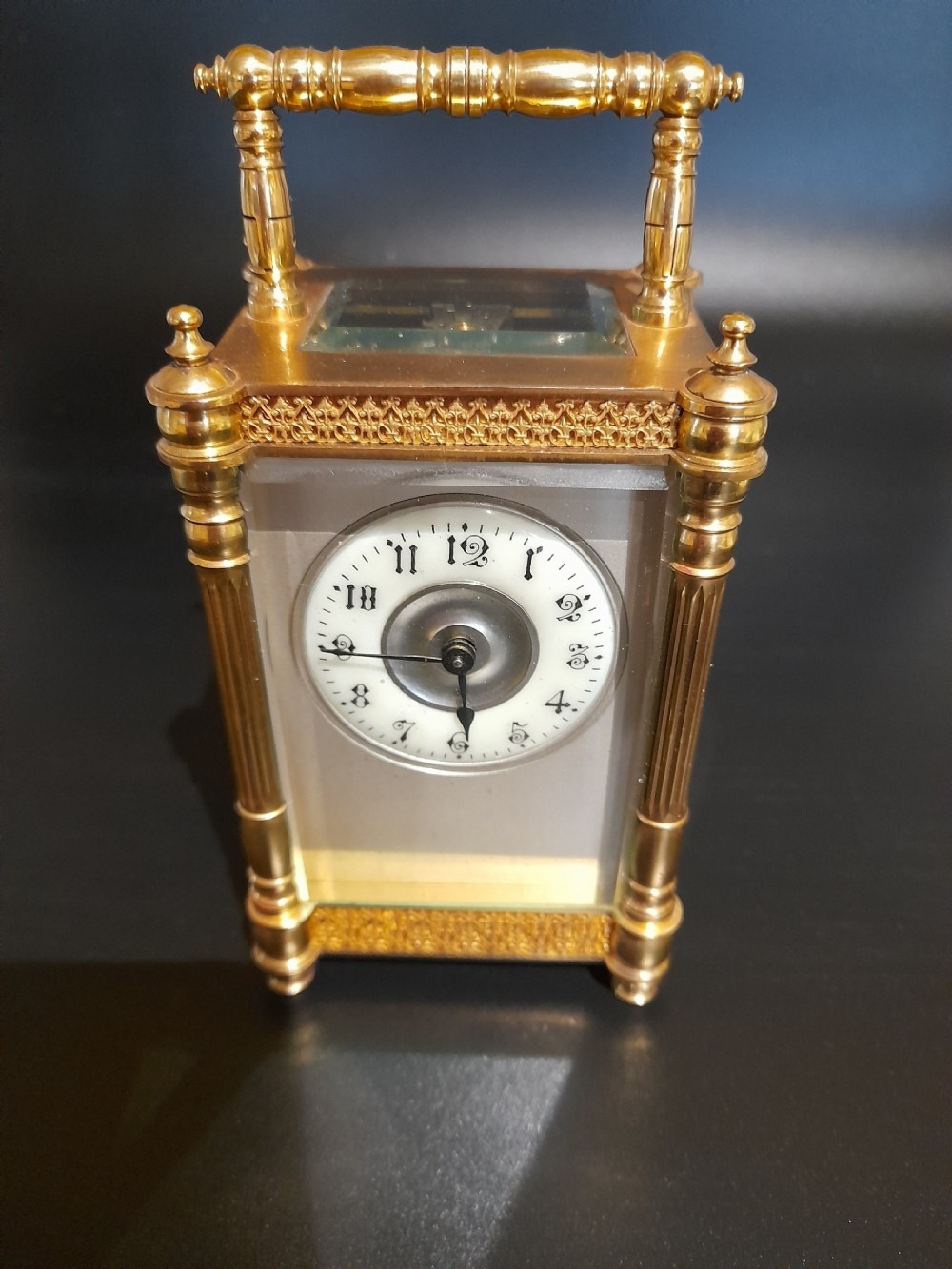 A gilded brass carriage clock featuring clear glass side panels, turned corner columns, a folding top handle, and a white enamel dial with Arabic numerals.