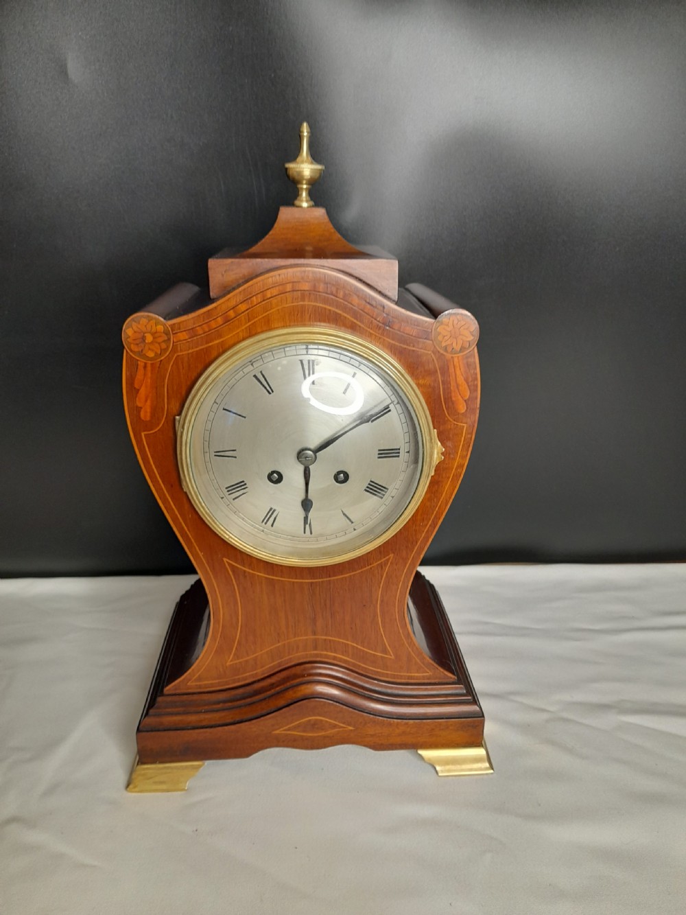 An Art Nouveau satinwood mantel clock featuring floral marquetry inlays, a silvered Roman numeral dial, a brass finial, and brass bracket feet.