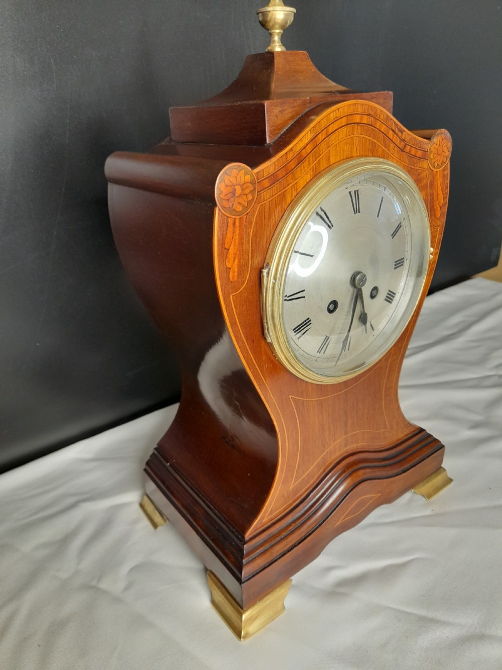 An Art Nouveau satinwood mantel clock featuring floral marquetry inlays, a silvered Roman numeral dial, a brass finial, and brass bracket feet. View V