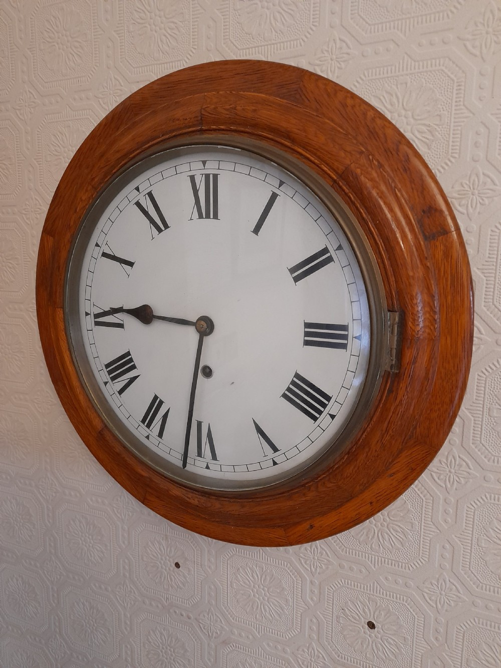 A circular antique golden oak wall clock featuring a white dial with black Roman numerals and a brass-rimmed glass cover.