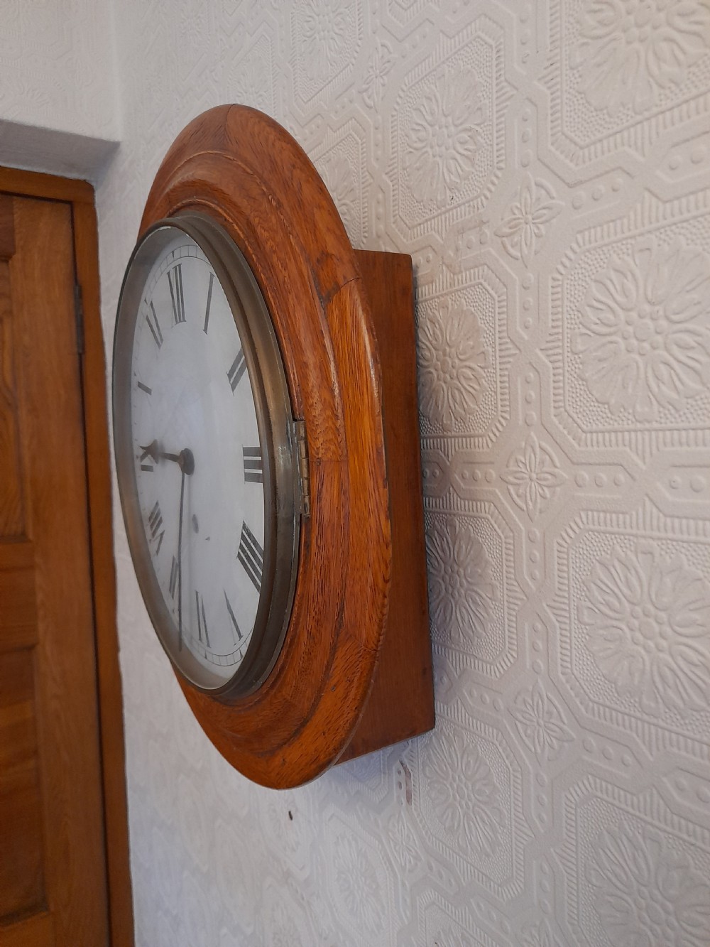 A circular antique golden oak wall clock featuring a white dial with black Roman numerals and a brass-rimmed glass cover. View III