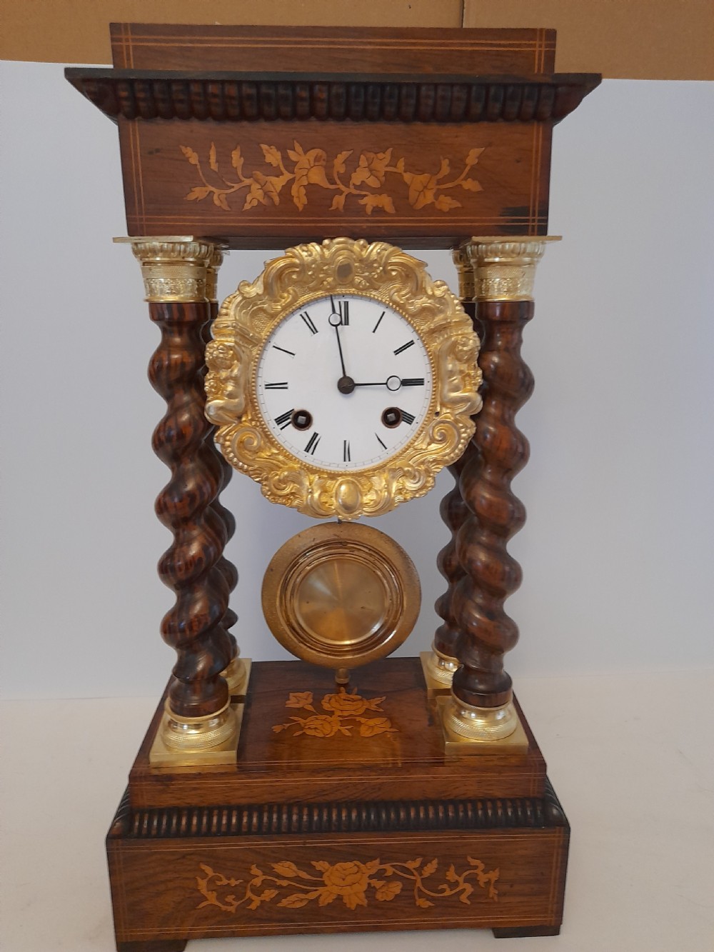 A 19th-century French rosewood portico clock featuring twisted columns, floral marquetry inlays, a white enamel dial with Roman numerals, and a gilt metal pendulum.