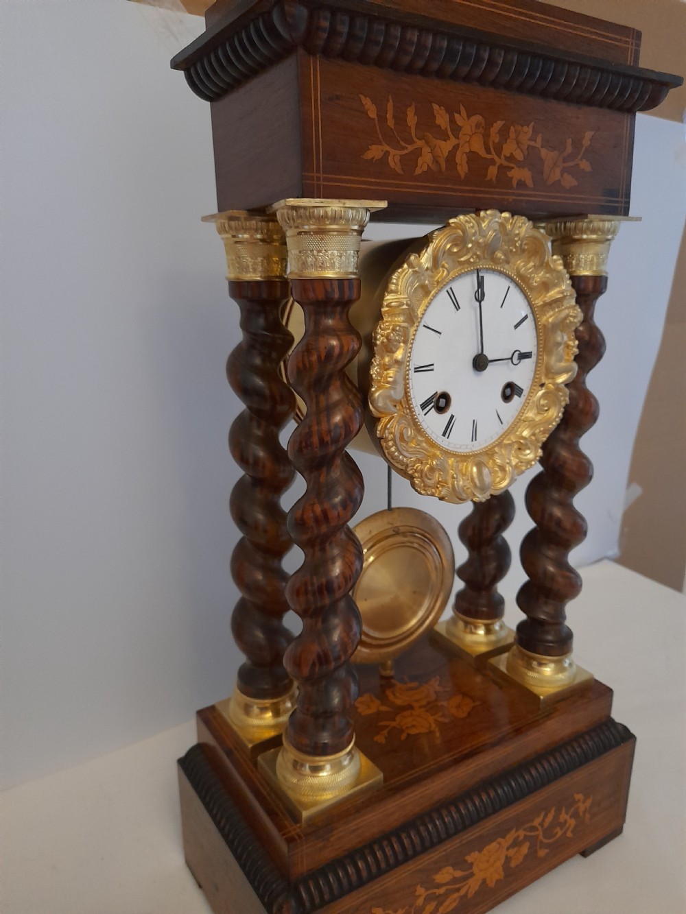 A 19th-century French rosewood portico clock featuring twisted columns, floral marquetry inlays, a white enamel dial with Roman numerals, and a gilt metal pendulum. View V
