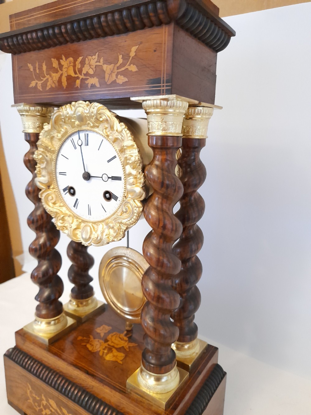 A 19th-century French rosewood portico clock featuring twisted columns, floral marquetry inlays, a white enamel dial with Roman numerals, and a gilt metal pendulum. View VI