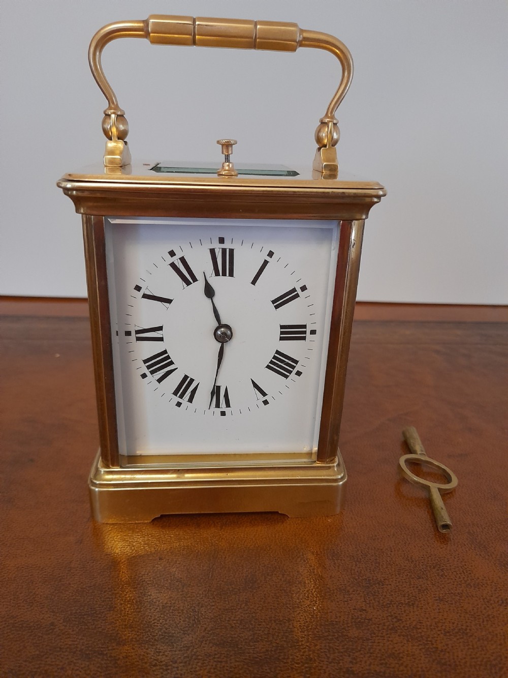 An antique brass repeating carriage clock featuring a white enamel dial with Roman numerals, bevelled glass panels, and a folding handle beside its key.