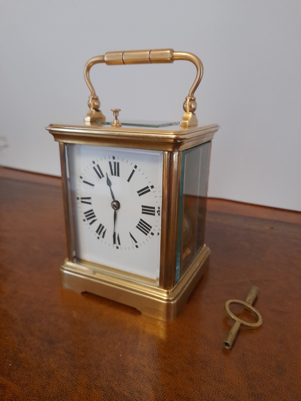 An antique brass repeating carriage clock featuring a white enamel dial with Roman numerals, bevelled glass panels, and a folding handle beside its key. View II
