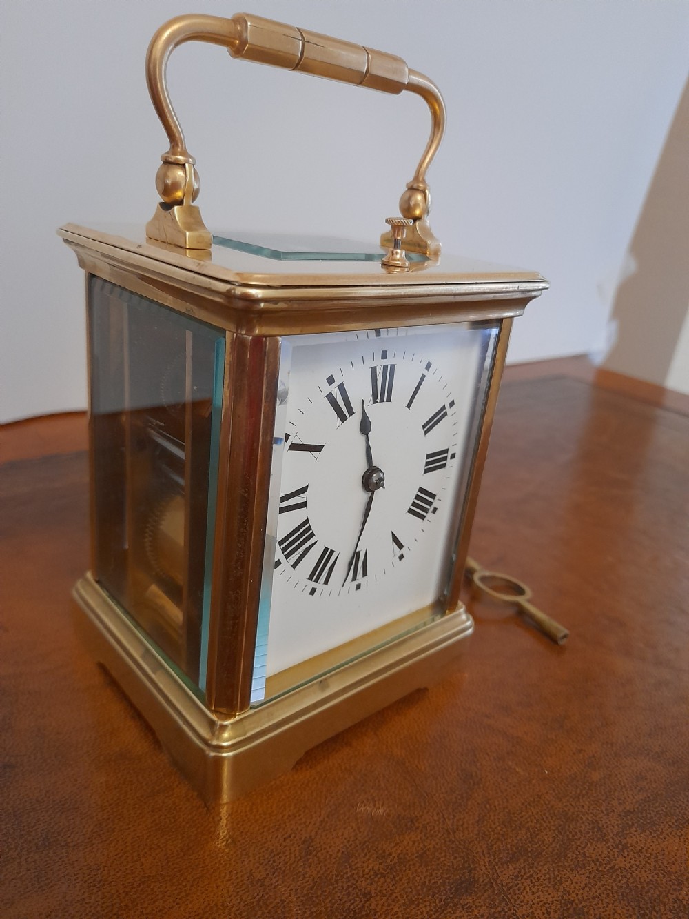 An antique brass repeating carriage clock featuring a white enamel dial with Roman numerals, bevelled glass panels, and a folding handle beside its key. View III