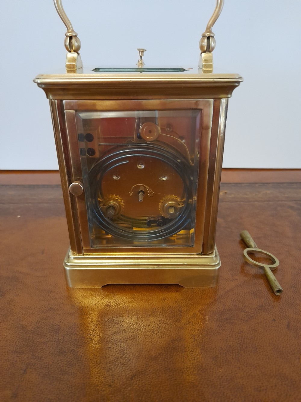 An antique brass repeating carriage clock featuring a white enamel dial with Roman numerals, bevelled glass panels, and a folding handle beside its key. View V
