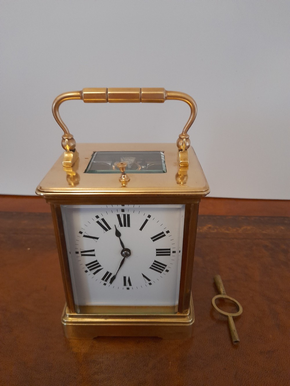An antique brass repeating carriage clock featuring a white enamel dial with Roman numerals, bevelled glass panels, and a folding handle beside its key. View VI