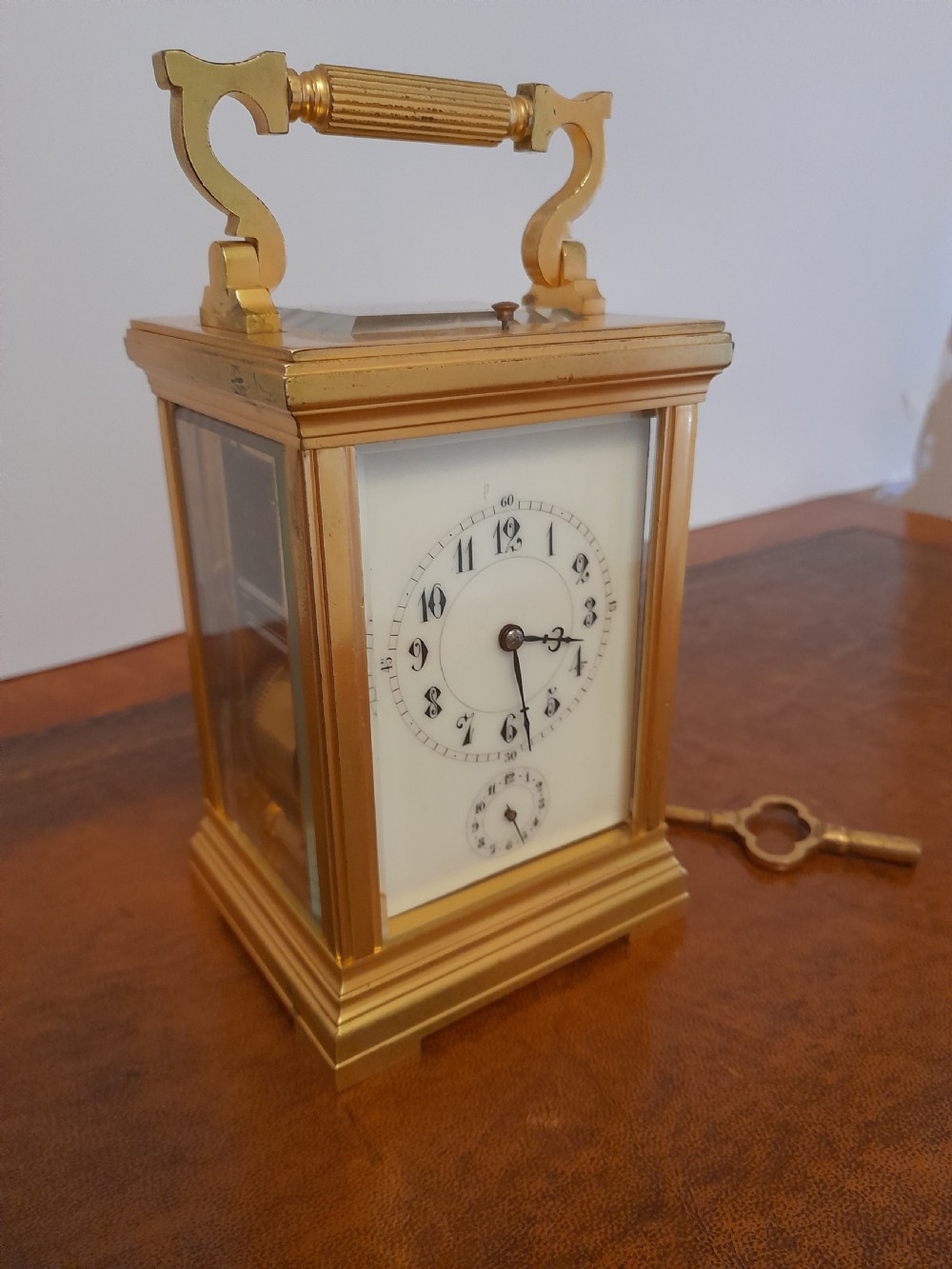 A gilt brass carriage clock featuring a fluted top handle, bevelled glass panels, a white enamel main dial with Arabic numerals, an alarm sub-dial, and an accompanying winding key. View II